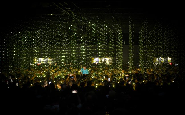 Four Tet stands in the centre of the crowd on a raised platform. He is wearing a blue t-shirt and faces the camera. Most of the audience have their backs to the camera, holding up phone screens to record video. Four Tet and the audience are surrounded by strands glowing in shades of green, which light up the faces of the audience closest to the centre.