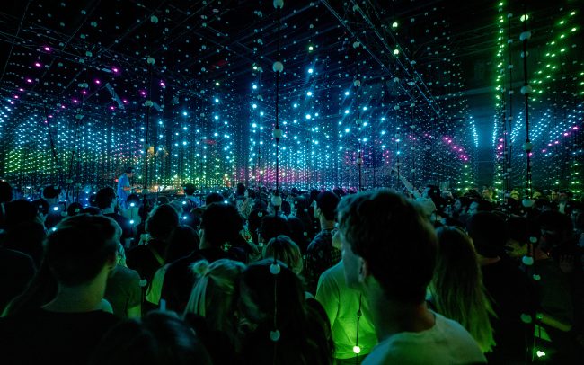 You are looking at Four Tet as though you are part of the crowd. He is in the distance and you are surrounded by vertically hanging LED strands, illuminated in a geometric pattern in shades of blue, green and pink. The people in front of you are lit up in bright green.