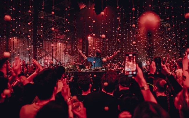 Four Tet stands at the centre of a large crowd, facing away from the camera with both hands raised to his side and palms facing the sky to signal the end of a show. The audience around him raise their hands above their head to clap. Both the audience and Four Tet are illuminated a deep red by the lights of the venue.